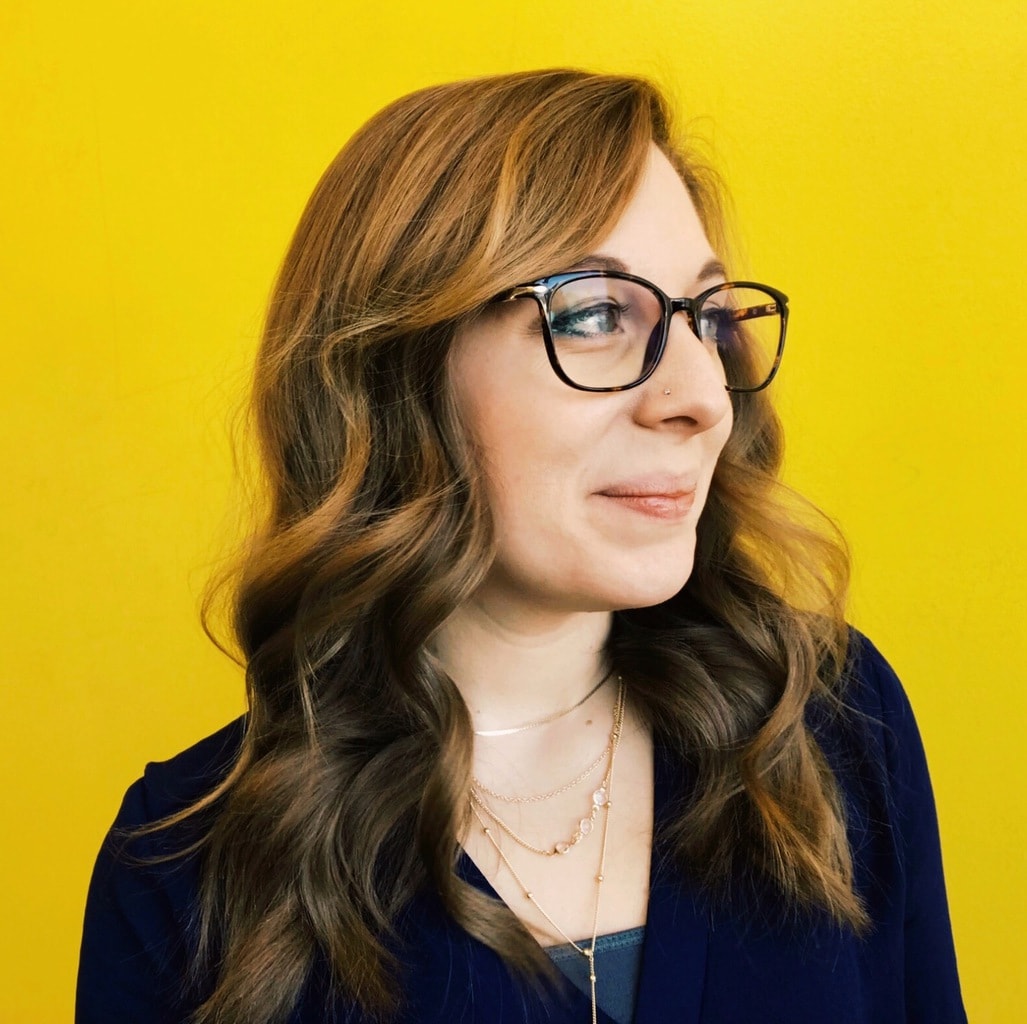 Brit Kolo headshot, a woman with long light brown hair, black glasses, and a yellow background.