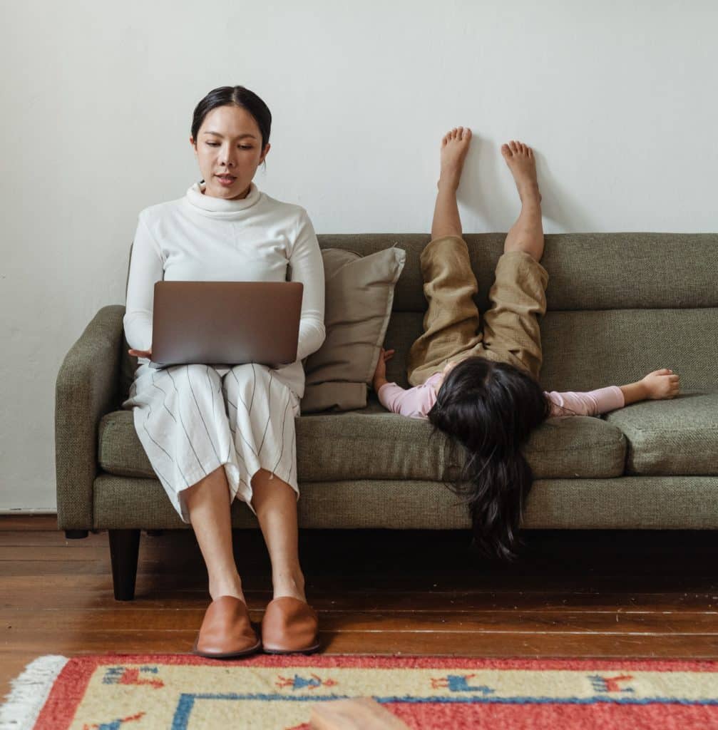A woman sits on a sofa, working on her laptop, while a girl lays on her back next to her. 