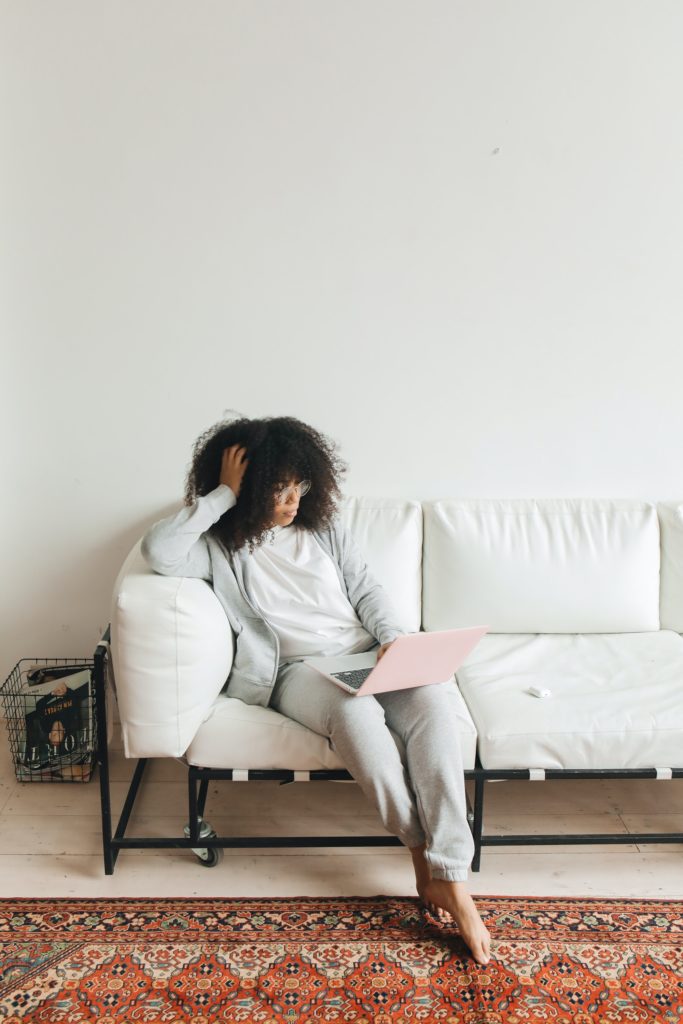 Woman with curly hair sits on a white couch, looking at her laptop. 